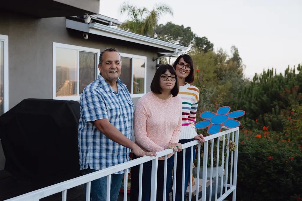 A family of three stands next to a white metal railing on the deck in the backyard of a house with trees around the exterior. The family is smiling and looking towards the camera, standing together.
