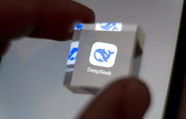 DeepSeek’s self The DeepSeek application icon displayed on a smartphone screen is magnified by a clear cube held between thumb and forefinger.