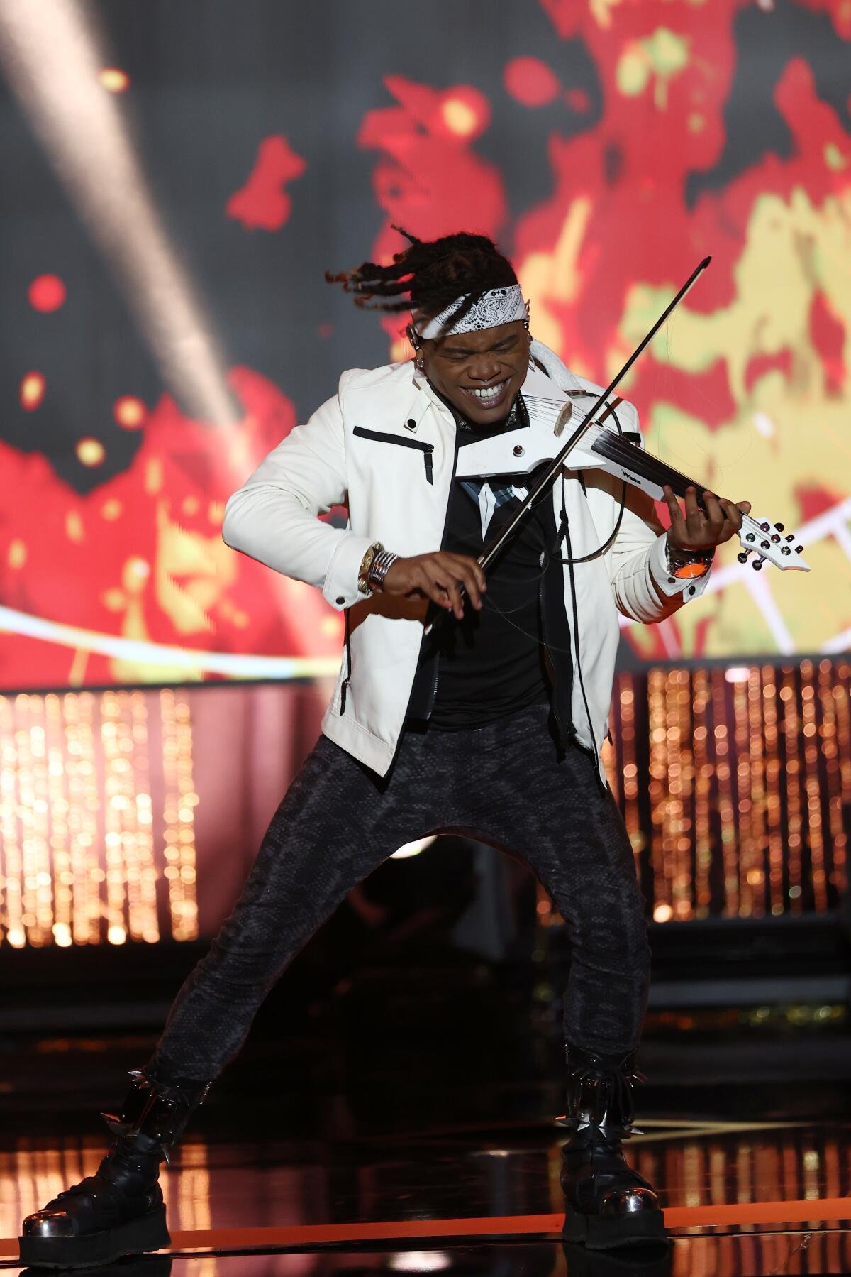 A man in black pants, a white jacket and white headband energetically playing an electric violin onstage.