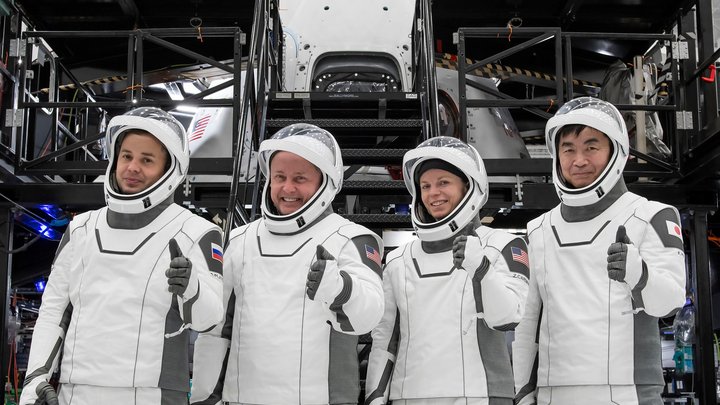 three men and one woman, all in white spacesuits, give a thumbs-up in a training facility here on earth